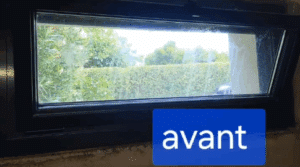 Fenêtre rectangulaire sombre vue de l'intérieur, donnant sur un jardin verdoyant avec une haie. Des marques sur la vitre. Texte "avant".