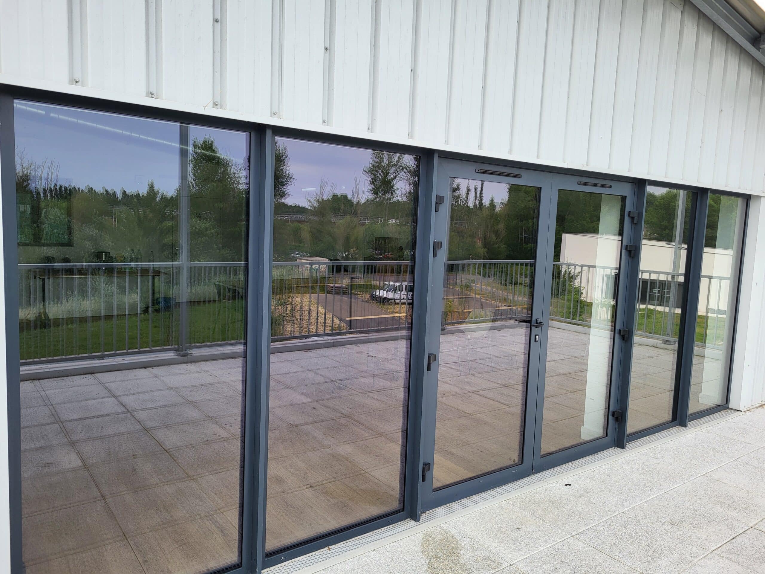 Façade moderne avec bardage blanc ondulé et grandes baies vitrées. Balcon carrelé gris clair reflétant arbres et véhicules. Portes vitrées anthracite.