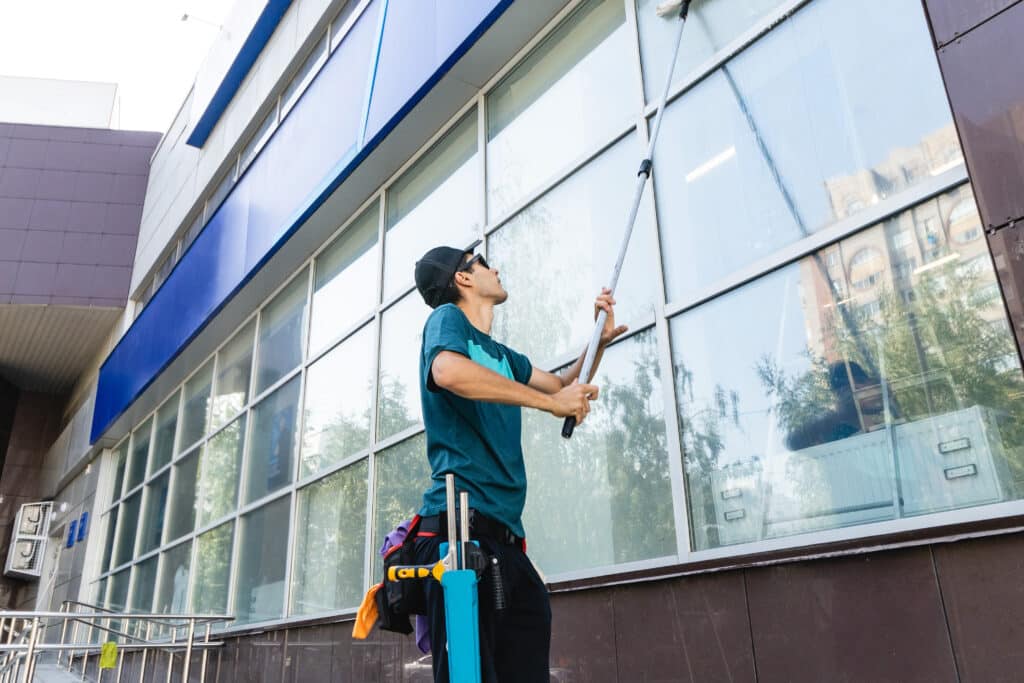 Jeune homme nettoie les grandes vitres d'un bâtiment moderne avec une raclette télescopique. Il porte une casquette, des lunettes de soleil et une ceinture d'outils.