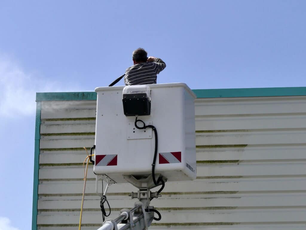 Un ouvrier en nacelle nettoie une façade de bâtiment couverte de verdure avec un nettoyeur haute pression, sous un ciel bleu dégagé.
