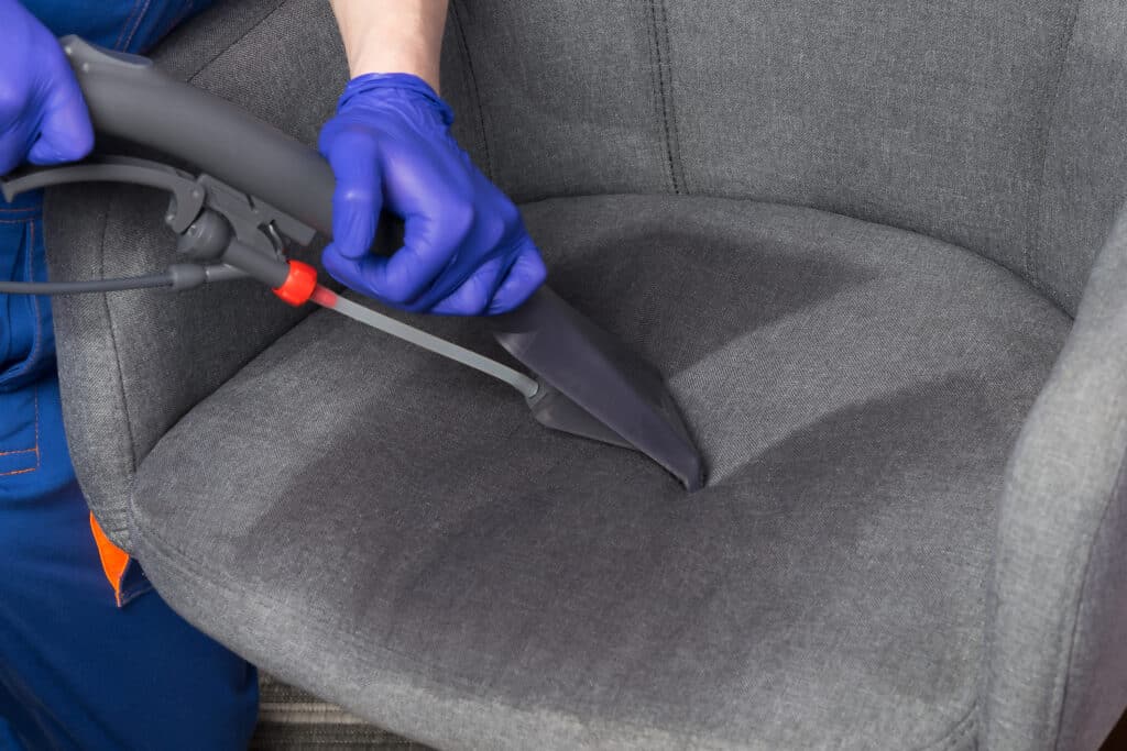 Personne en gants bleus nettoyant en profondeur un fauteuil gris en tissu avec un extracteur professionnel, éliminant saleté et taches.