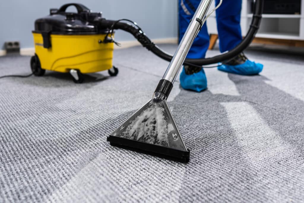 Un professionnel nettoie un tapis gris à motifs avec une machine d'extraction, laissant une bande propre sans mousse ni saleté.