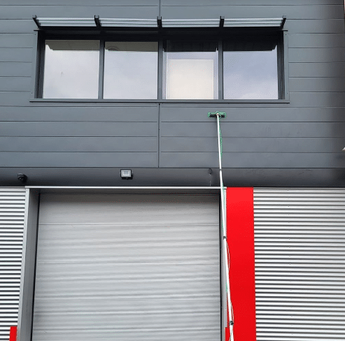 Façade de bâtiment gris avec fenêtres, porte de garage et perche télescopique verte pour le nettoyage des vitres.