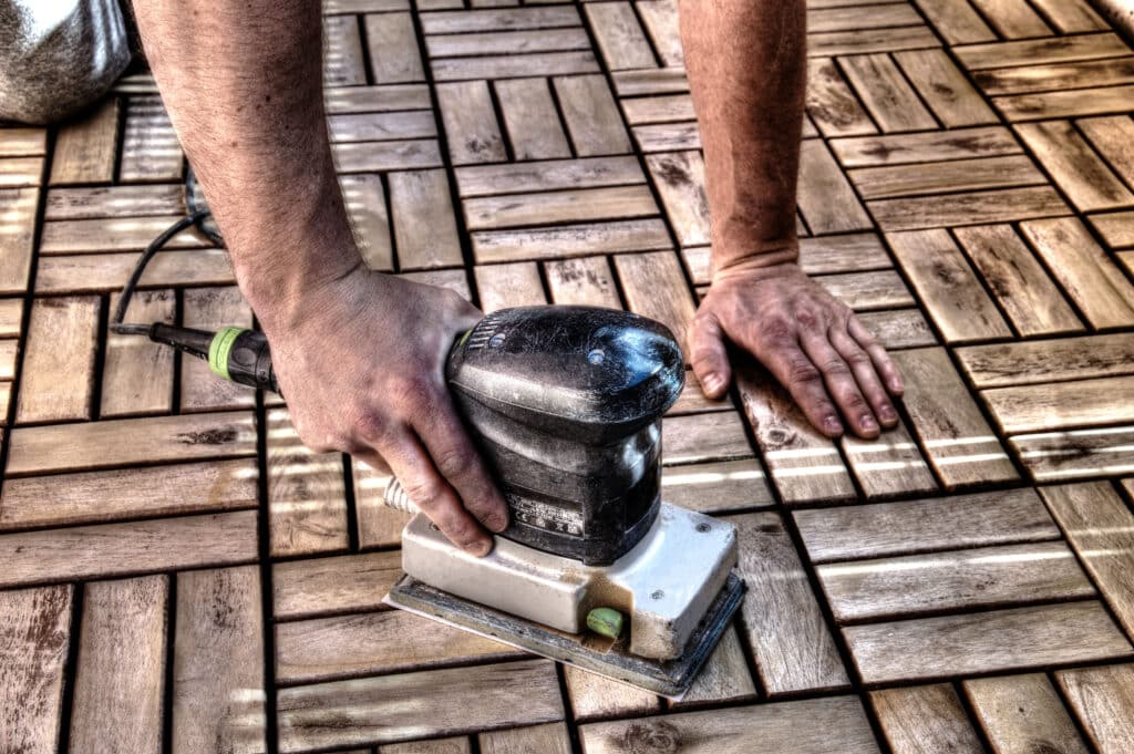 Main ponçant des dalles de terrasse en bois avec une ponceuse orbitale. Le bois est usé, le ponçage est en cours.