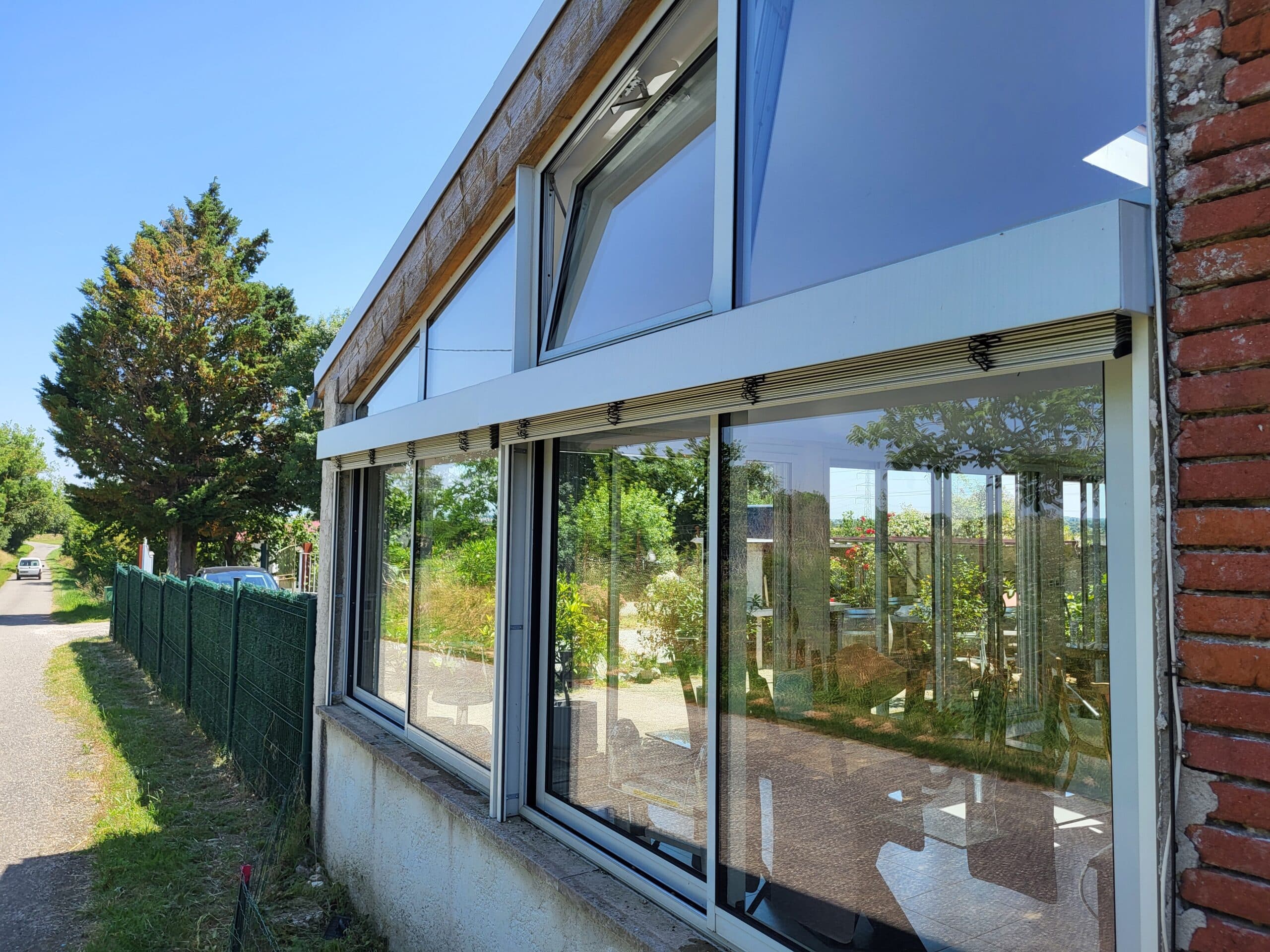Véranda moderne vitrée avec charpente bois claire et mur de briques rouges. Les grandes fenêtres reflètent route, arbres verts et ciel bleu.