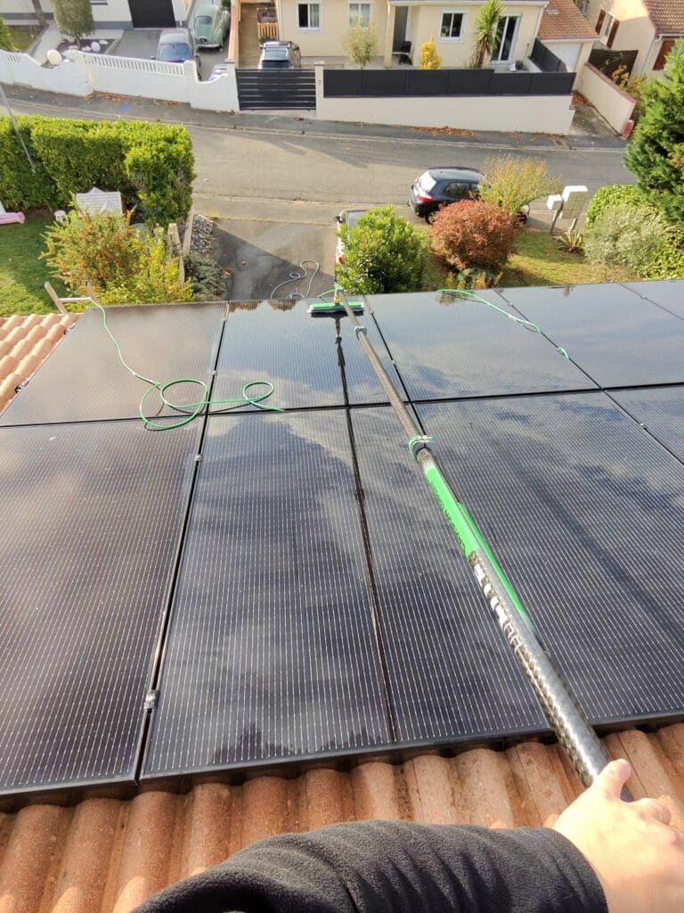 Nettoyage de panneaux solaires noirs sur toit en tuiles avec une perche télescopique munie d'une brosse et un tuyau vert, vue d'un quartier résidentiel en contrebas.