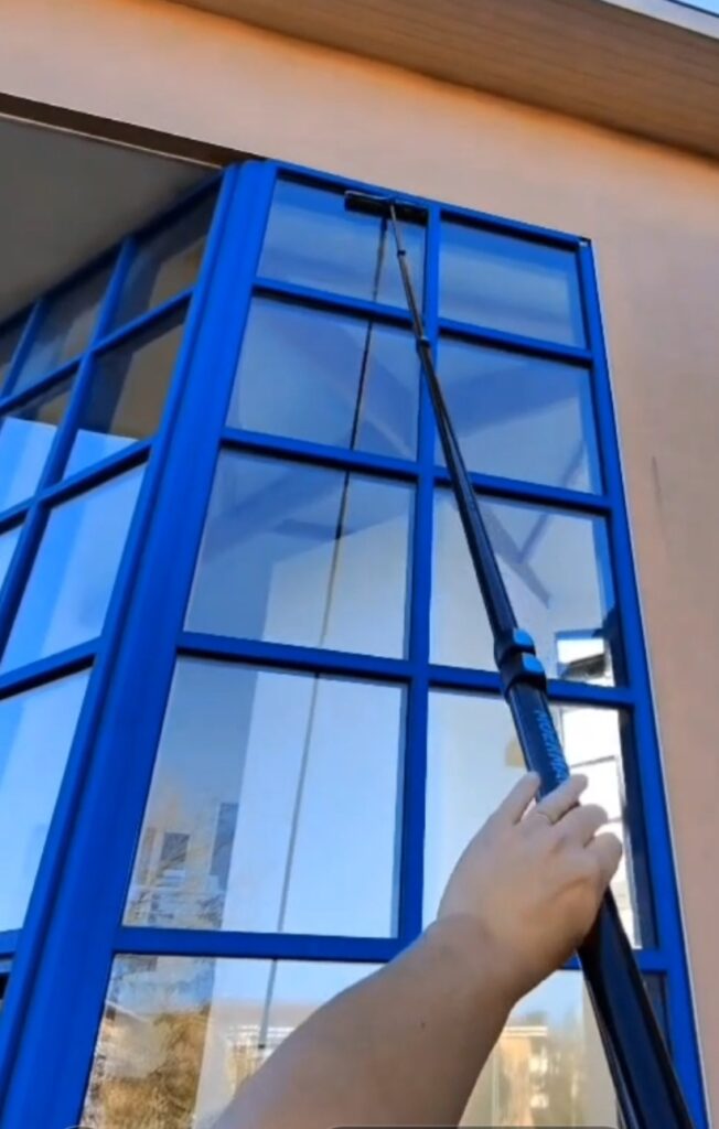 Nettoyage de façade. Une main dirige la perche télescopique Moerman pour racler une grande vitre encadrée de bleu.