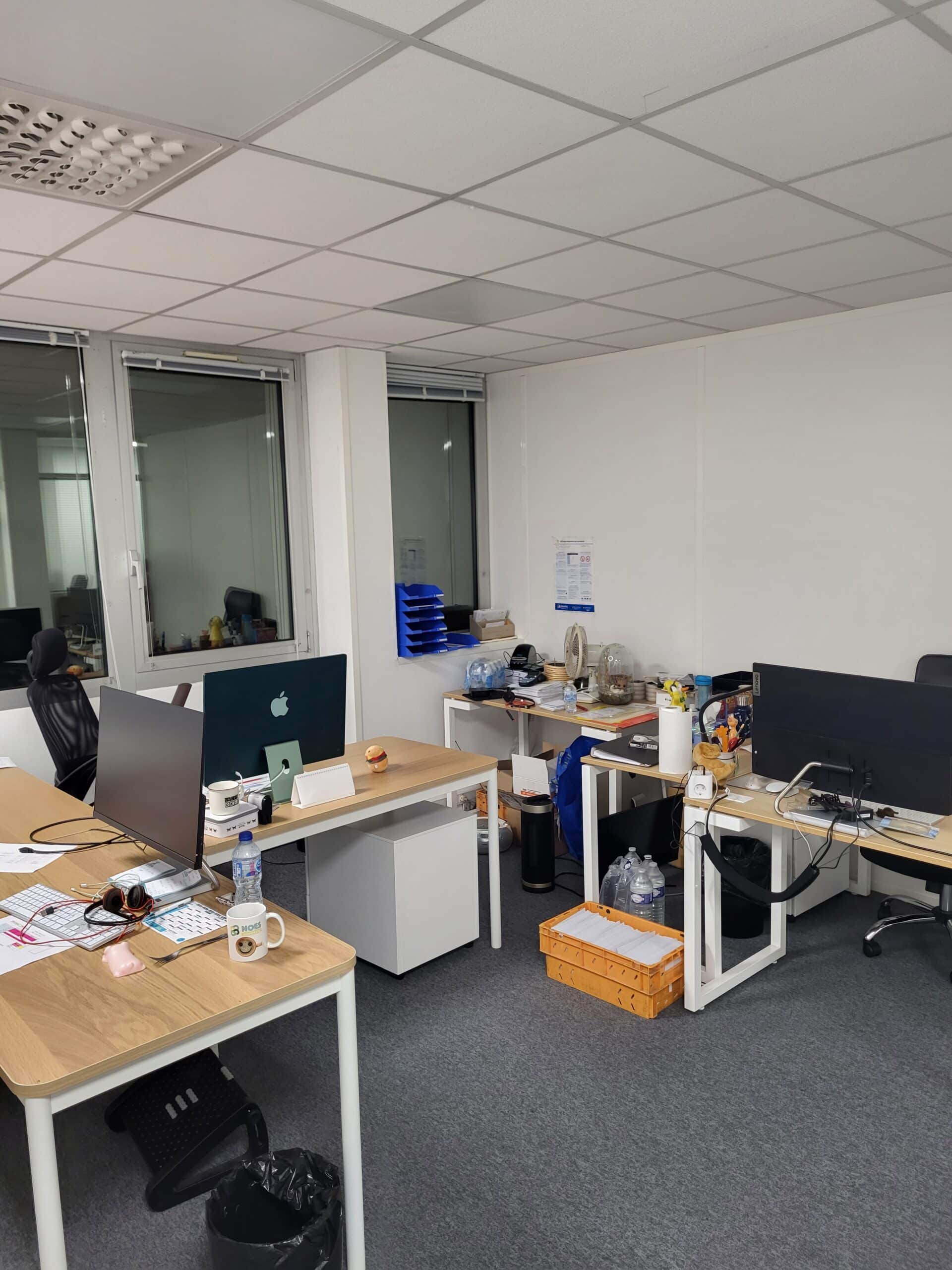 Espace de bureau moderne et fonctionnel (Apple/PC). Coin bureau moderne avec deux postes de travail encombrés, écrans Apple et PC, et fenêtres donnant sur un couloir. Mobilier clair, tapis gris.