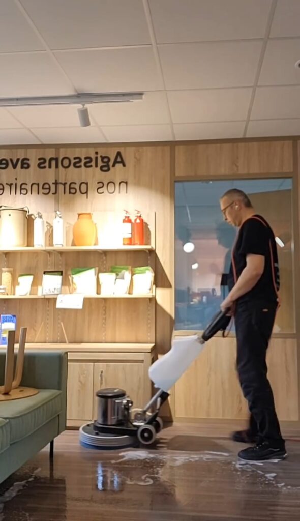 Homme utilisant une machine monobrosse pour le nettoyage et le lustrage d'un sol en bois dans un magasin. Le texte mural est «svA zossigA».