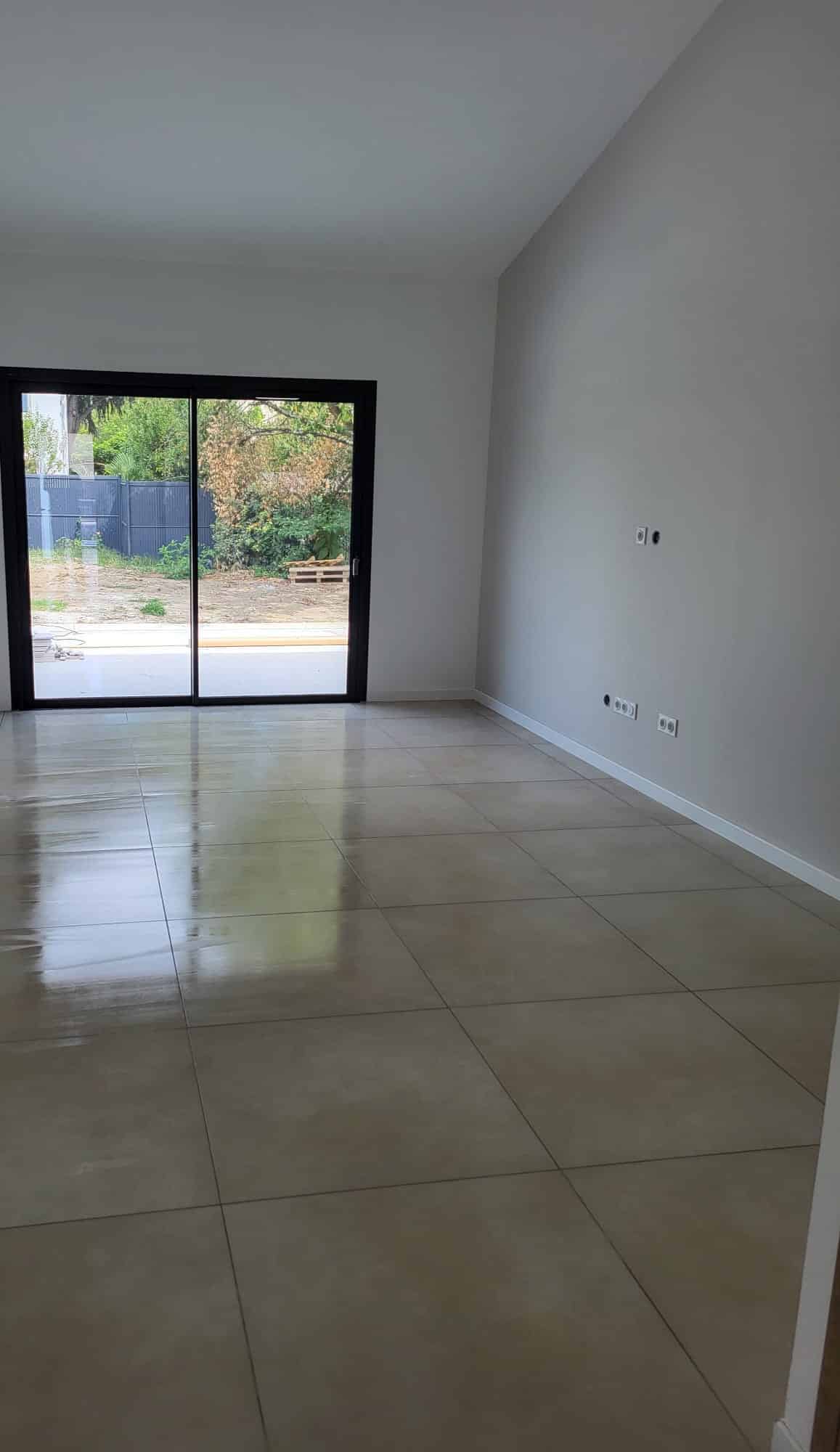 Salon neuf moderne : carrelage brillant et baie vitrée Pièce de vie moderne vide, murs blancs, grand carrelage beige brillant. Baie vitrée coulissante noire et prises électriques sur le mur droit.