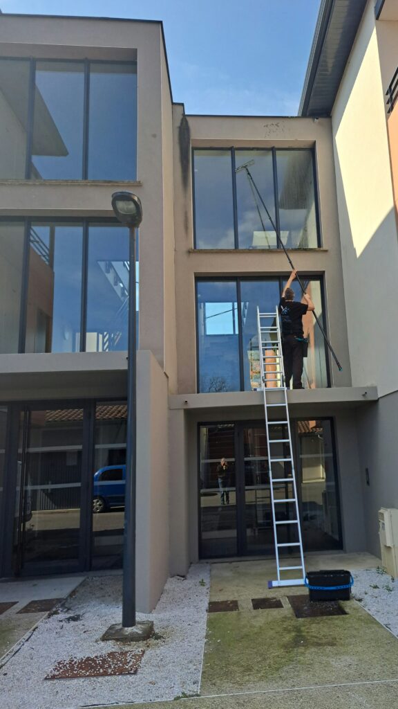 Laveur de vitres professionnel utilisant une perche télescopique sur une échelle pour nettoyer un immeuble moderne.